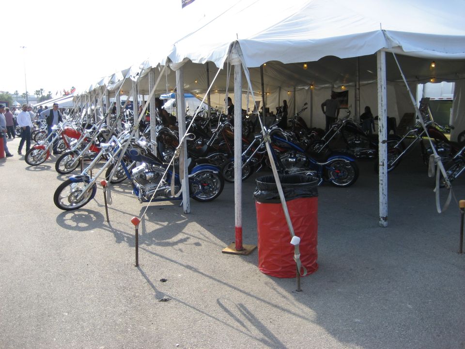 Multiple custom chopper motorcycles parked in a row under a white tent. Some bikes are red, blue, and black