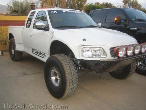 1990 Ford Bronco - white lifted Ford prerunner pickup displays a row of round driving lights, BFGoodrich decals, and exposed long-travel front suspension.