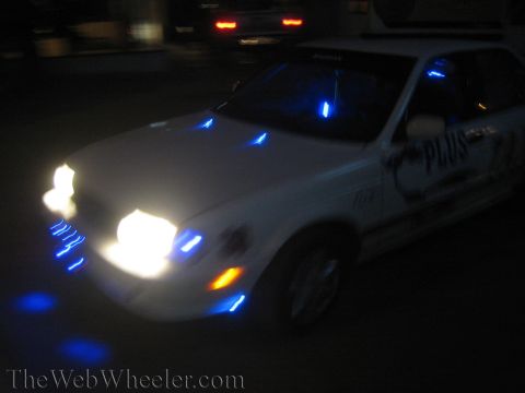 1990 Ford Bronco - white car cruises at night covered in glowing blue and orange LED accent lights, captured in a blurred low-light shot.
