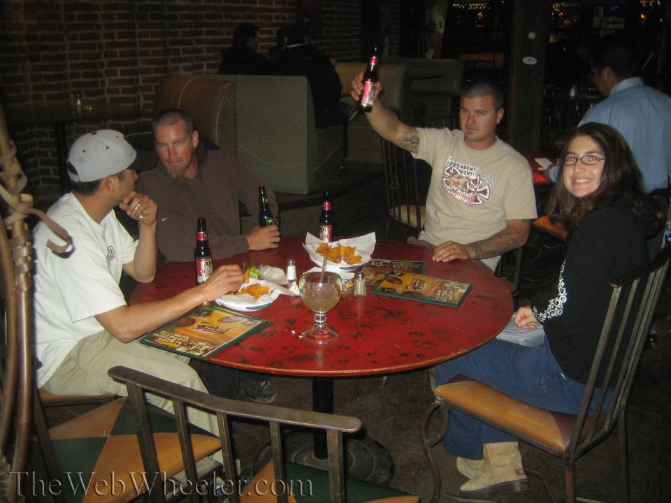 Four people seated around a round red table with beer bottles and snacks, one raising a bottle