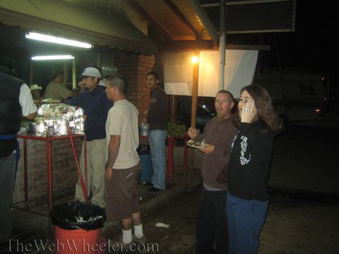 1990 Ford Bronco - People gather at an outdoor food stand at night under a brick overhang, two of them holding plates beside a steam-table cart.