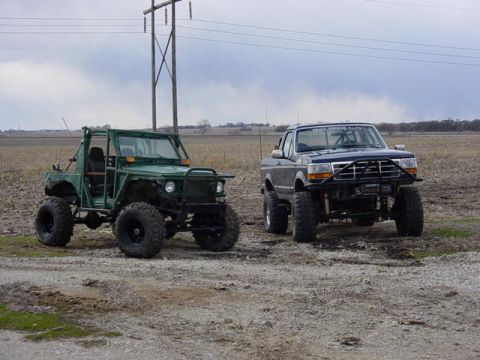 1992 Ford Bronco - Two modified off-road vehicles are parked on a dirt path under a cloudy sky.