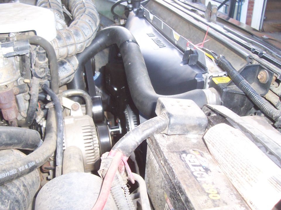 Engine bay view showing radiator, fan, alternator, and battery. Radiator has text that is difficult to read
