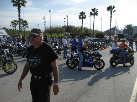 1986 Ford Bronco - A crowd gathers around parked motorcycles under a bright, sunny sky with palm trees and a stadium visible.