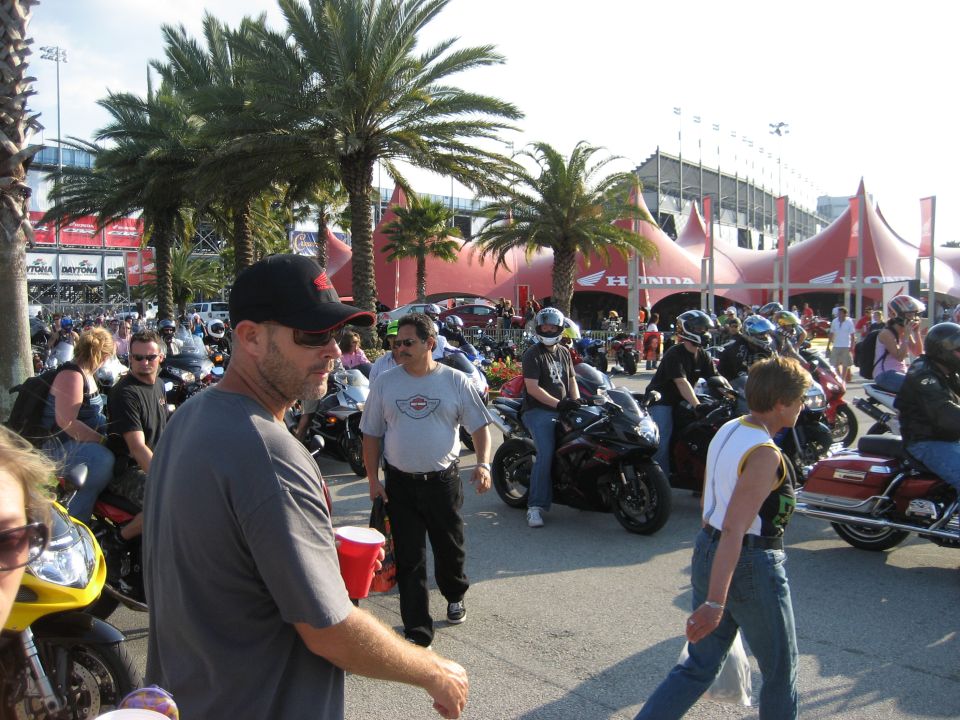 Crowd of people and many motorcycles gathered outdoors with palm trees and a large tent structure in the background