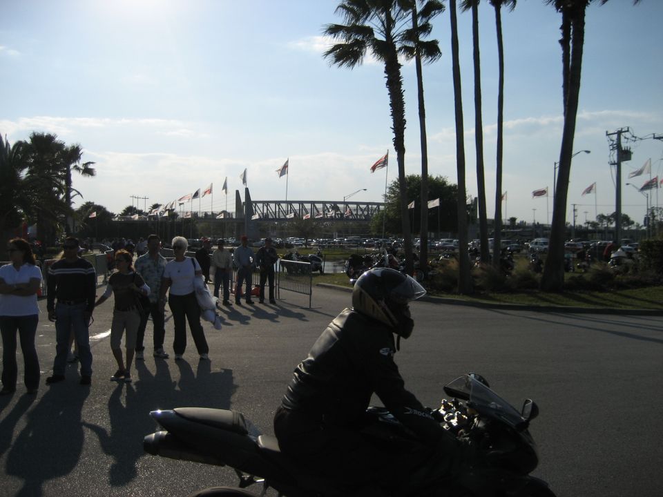 Motorcycle rider in foreground, crowd of people walking in background with palm trees and a bridge