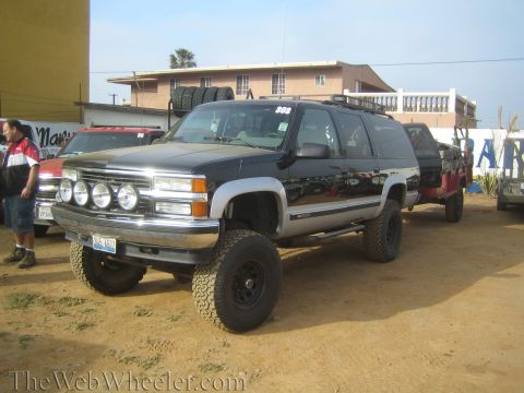 1990 Ford Bronco - black lifted Chevrolet Suburban with chrome bumper and four round driving lights tows a trailer across a dirt lot near buildings.