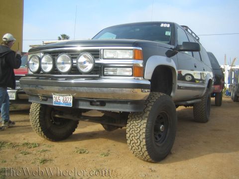 1990 Ford Bronco - front of a black lifted Chevrolet SUV shows a chrome bumper, four round driving lights, and large off-road tires on a dirt lot.