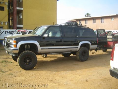 1990 Ford Bronco - Side view of a black lifted Chevrolet Suburban with a roof rack and chunky off-road tires parked on dirt among other vehicles.