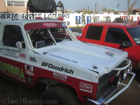 1990 Ford Bronco - red-and-white off-road race truck plastered with BFGoodrich, Bilstein, and California Tomatoes sponsor decals sits beside a red SUV.