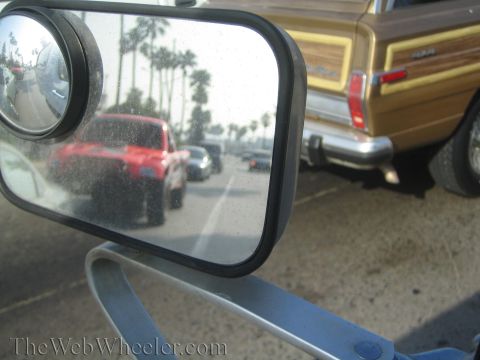 1990 Ford Bronco - convex blind-spot mirror reflects street traffic and palm trees while a brown wood-panel station wagon sits to the right.