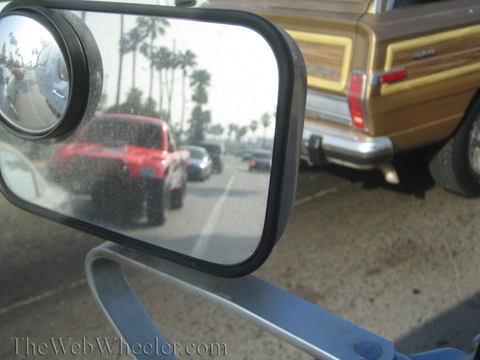 Convex blind-spot mirror reflecting traffic and palm trees, with a brown woody wagon at right