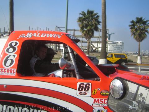 1990 Ford Bronco - red off-road race truck numbered 86 and 66 with a driver aboard moves along a road past a harbor where a cruise ship is docked.