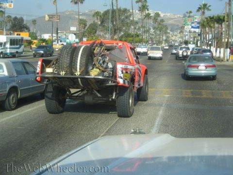 1990 Ford Bronco - rear of an orange open-cockpit off-road race buggy with spare tires sits in street traffic beneath palm-lined hills.