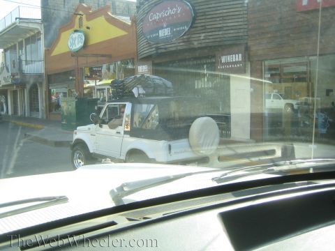 1990 Ford Bronco - white compact SUV with a roof rack and a bed-mounted spare tire is parked on a town street, viewed through a car windshield.