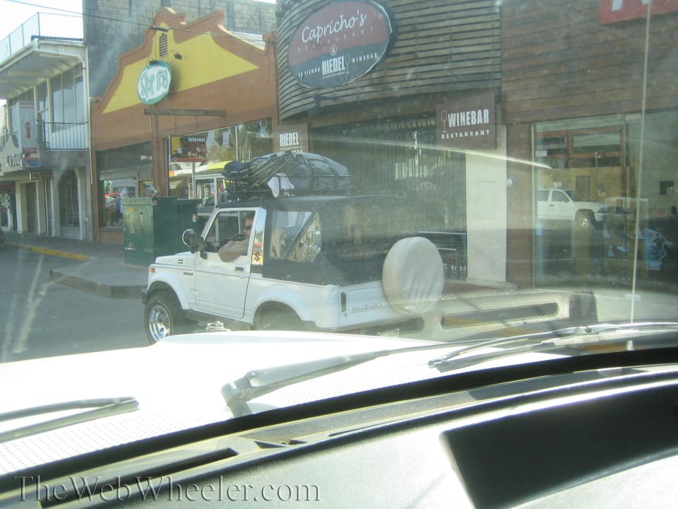 White small SUV with roof rack and bed-mounted spare on a town street, seen through a windshield