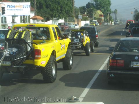 1990 Ford Bronco - yellow off-road pickup with bed-mounted spare tires tows a red race buggy through street traffic past a Pablo Alejo 2007 banner.