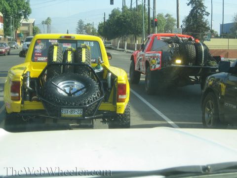 1990 Ford Bronco - rear of a yellow off-road pickup loaded with spare tires tows a red race truck on a flatbed through city traffic.