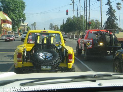 1990 Ford Bronco - yellow off-road pickup with bed-mounted spare tires tows a red race truck on a trailer at a stoplight along a palm-lined road.