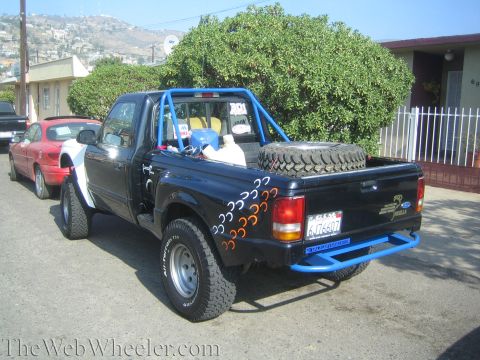 1990 Ford Bronco - Rear-quarter view of a black Ford Ranger prerunner pickup with a blue exterior roll cage, flame graphics and a spare tire in the bed.