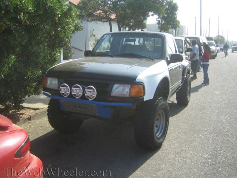 1990 Ford Bronco - Front-quarter view of a lifted black-and-white Ford Ranger prerunner with three Hella lights on a blue bumper, parked on a street.
