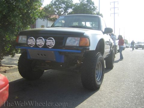 1990 Ford Bronco - Front view of a lifted black-and-white Ford prerunner pickup on large tires with three Hella lights mounted on a blue front bumper.