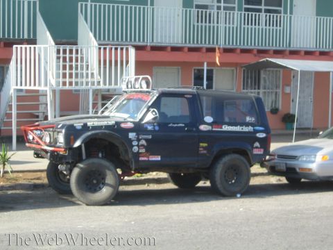 1990 Ford Bronco - Side view of a dark blue lifted two-door off-road truck covered in BFGoodrich and sponsor decals, parked before a pink building.