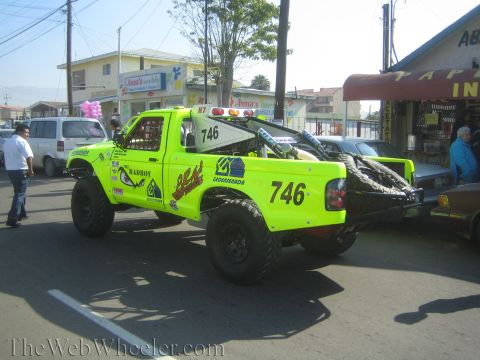 1990 Ford Bronco - Rear-quarter view of a bright green off-road race truck numbered 746 with sponsor decals, parked on a street near shops.