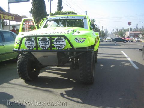 1990 Ford Bronco - Head-on view of a bright green off-road race truck with three Hella lights and a tube bumper driving on a road.