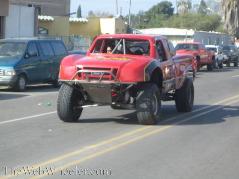 1990 Ford Bronco - Front-quarter view of a red off-road race truck with a Ford grille emblem and tube bumper on large tires driving down a street.