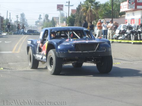 1990 Ford Bronco - Front-quarter view of a dark blue off-road race truck with Red Bull livery and a tube bumper, with spectators and motorcycles behind.