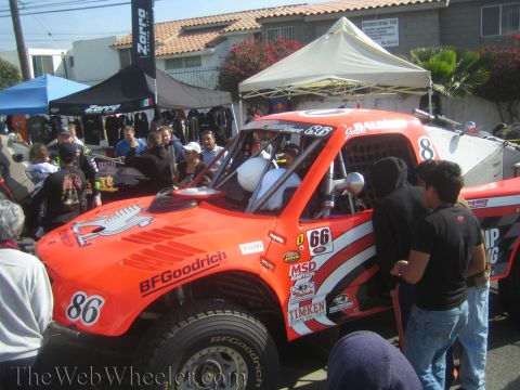 1990 Ford Bronco - Orange-and-white off-road trophy truck numbered 86 with BFGoodrich decals surrounded by people in a pit paddock under canopies.
