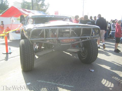 1990 Ford Bronco - Front view of a black off-road trophy truck showing its tube-frame front suspension, parked among a crowd of spectators.