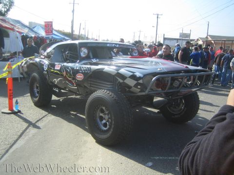 1990 Ford Bronco - Front-quarter view of a black off-road race truck with checkered-flag and flame graphics on large tires amid a street crowd.