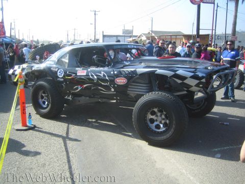 1990 Ford Bronco - Side view of a black off-road race truck with checkered-flag and flame graphics and large off-road tires among spectators.
