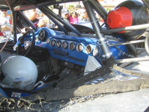 1990 Ford Bronco - Cockpit view of a blue off-road race truck with a row of round gauges, steering wheel, helmet and roll-cage tubing.