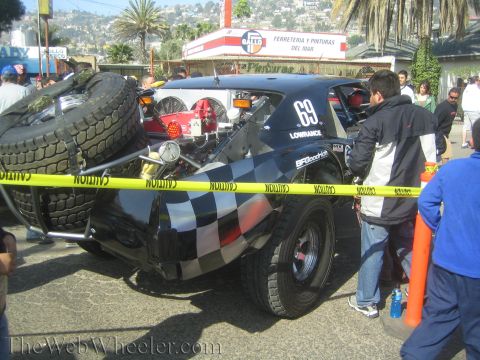 1990 Ford Bronco - Rear-quarter view of a black checkered off-road race truck numbered 69 with a spare tire on the back, behind yellow caution tape.
