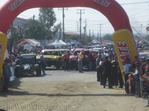 1990 Ford Bronco - Off-road race trucks staged on a dirt road beneath a red-and-yellow Tecate inflatable start arch with crowds lining the sides.