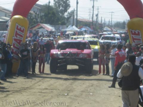 1990 Ford Bronco - Head-on view of a red off-road race truck at the start line under a Tecate arch, flanked by spectators and grid girls.