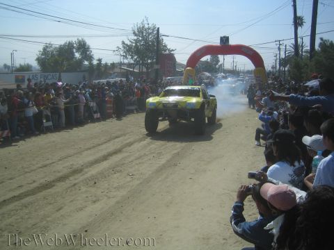 1990 Ford Bronco - Yellow off-road race truck launching off a dirt start line in a cloud of dust as a large crowd watches from behind barriers.