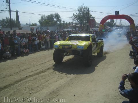 1990 Ford Bronco - Yellow off-road race truck accelerating down a dirt course kicking up dust, with a crowd and inflatable arch in the background.