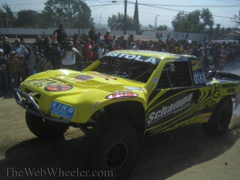 1990 Ford Bronco - bright yellow off-road trophy truck with Schampa and Precision Gear sponsor decals sits on a dirt course as spectators line a fence behind…