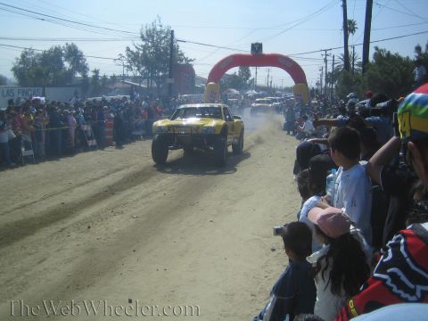 1990 Ford Bronco - yellow off-road race truck kicks up dust racing down a dirt road toward a red Lucas inflatable arch as crowds watch from both sides.