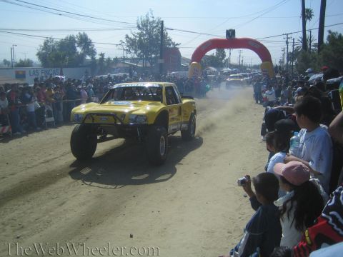 1990 Ford Bronco - yellow Ford trophy truck rolls along a dirt course lined with spectators, with a red inflatable race arch in the background.