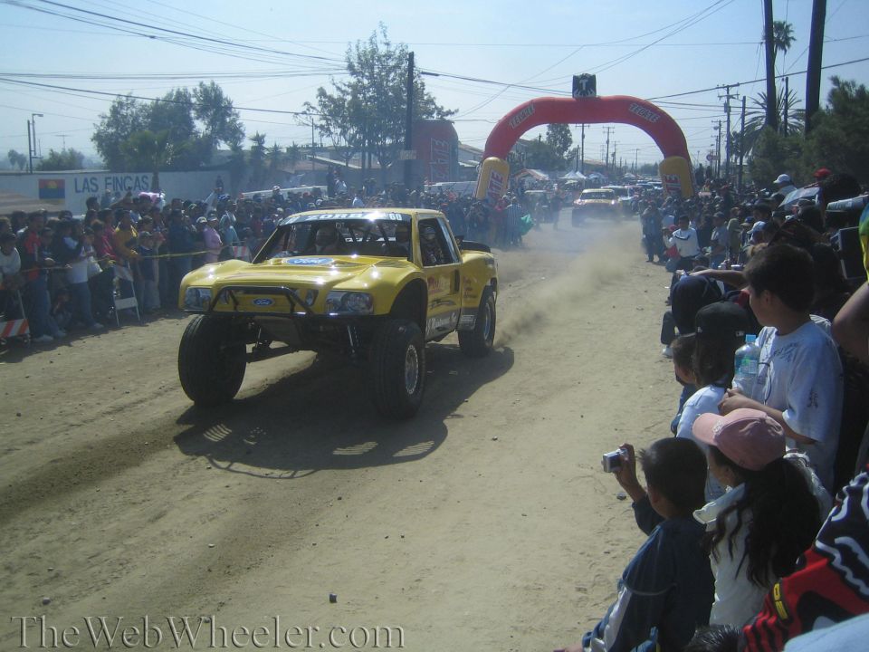 Yellow Ford off-road trophy truck on dirt course with spectators and inflatable arch
