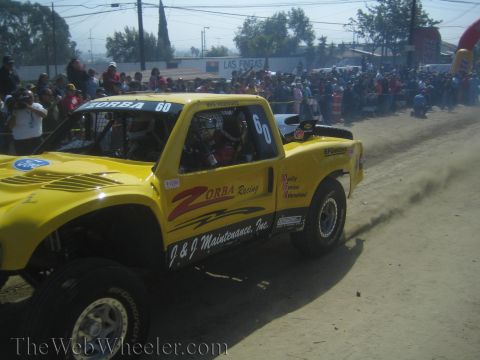 1990 Ford Bronco - yellow Zorba Racing off-road truck marked number 60 drives along a dusty course as a crowd of spectators looks on.