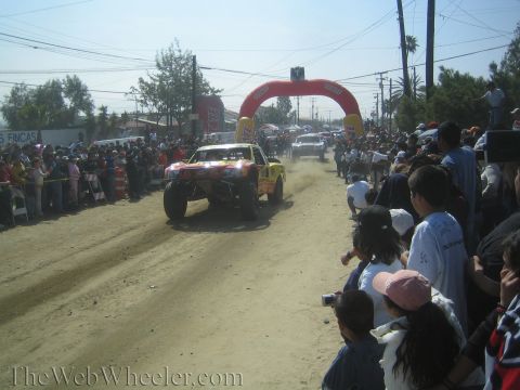 1990 Ford Bronco - off-road trophy truck speeds down a dusty dirt road toward a red inflatable arch with spectators crowding both sides.