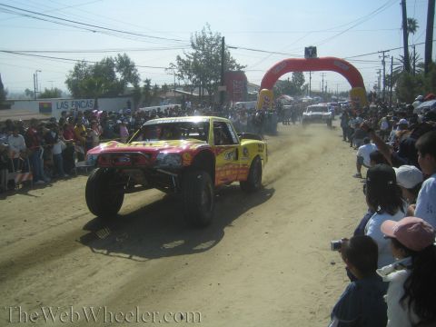 1990 Ford Bronco - pink and yellow off-road trophy truck catches air on a dirt course as crowds watch near a red inflatable race arch.