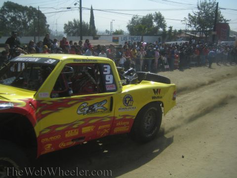 1990 Ford Bronco - yellow off-road truck with flame graphics and G&R Racing decals, numbered 39, kicks up dust before a packed crowd.