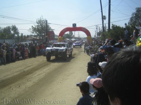 1990 Ford Bronco - white off-road trophy truck drives down a dusty dirt road toward a red inflatable arch with dense crowds on both sides.